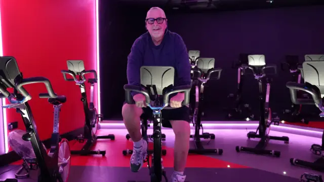 Kısa beyaz saçlı ve sakallı orta yaşlı bir adam olan Steve, bir spor salonunda bisiklet üzerinde egzersiz yapıyor. Pedalları çevirirken yüzünde bir gülümsemeyle kameraya bakıyor. Oda pembe ve mor renklerle aydınlatılmış.