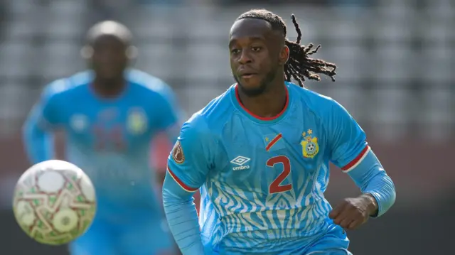 Aaron-Wan Bissaka, wearing a light blue DR Congo shirt with a red number two on the chest, is seen running as he chases a ball that is out of focus in the foreground. A DR Congo team-mate is seen in the background out of focus.