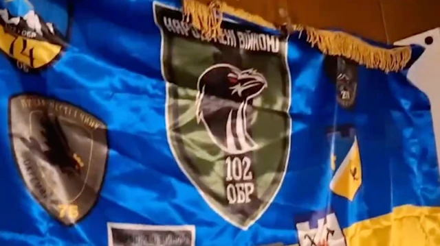 Синьо-жовтий прапор із жовтою бахромою по верхньому краю. На прапорі розміщено кілька емблем військових підрозділів. Центральна — великий зелений щит із чорною птахою та написом "Народжені війною" і "102 ОБР". Інші нашивки мають різні форми та кольори, зображення гір, птахів і символів, а також цифри "76" і "75".