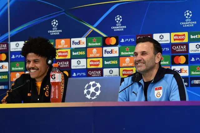 Gabriel Sara ve Okan Buruk Liverpool maçı öncesinde basın toplantısında görülüyor. Gazetecilerin sorularını yanıtlayan ikili, gülümsüyor.