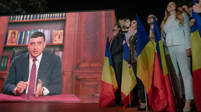 Bir ekranda George Simion, yanında Romanya bayrağı tutan kadın ve erkekler