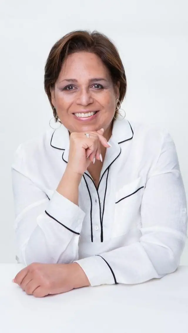 Fracis Bell, vestida con una blusa blanca, ve a la cámara mientras se sostiene el mentón con una de sus manos.
