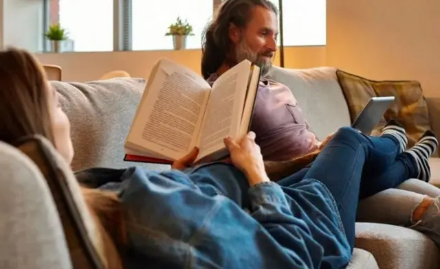 A girl is lying on the couch with a book in her hand and her legs in the lap of a man.