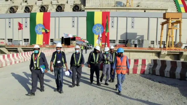 Un grupo de trabajadores de construcción caminan con cascos y tapabocas, mientras detrás de ellos se ve la bandera etíope ondeando en una sección de la Gran Presa del Renacimiento Etíope, en Benishangul-Gumuz, Etiopía.