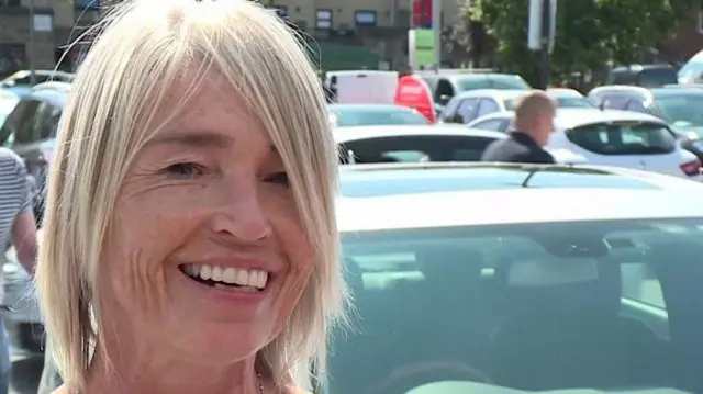 駐車場で車の前に立つキルナーさん。金髪で、笑顔を浮かべている