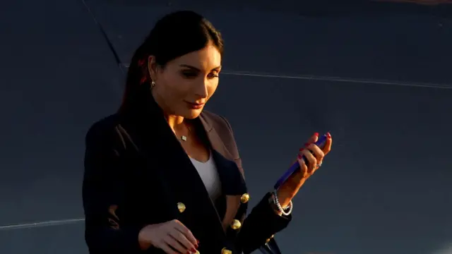 Kobieta o ciemnych włosach, ubrana w ciemną marynarkę, pod którą widać biały t-shirt, patrzy w dół przed siebie, trzymając w lewej dłoni telefon