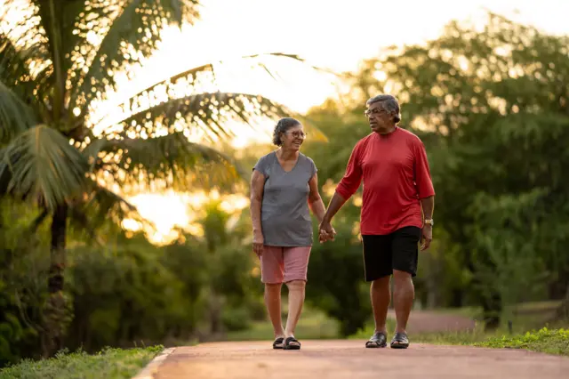 Um homem e uma mulher idosos caminham, de bermudas e camisetas, em uma área asfaltada de um parque