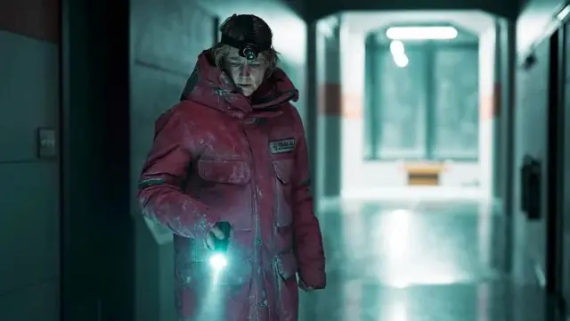 Jodie Foster é uma atriz loira, de pele clara e, no papel de Liz Danvers, está vestida com um casaco vermelho, em um corredor escuro e leva uma lanterna na mão direita apontada para o chão, e outra na testa.