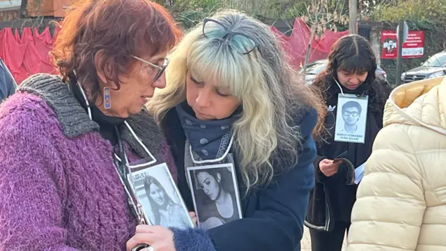 Holzapfel, à gauche, est vue avec une autre femme blonde à sa droite. Les femmes se trouvent à l'extérieur du site commémoratif, portant autour du cou des photos commémoratives en noir et blanc de personnes disparues et d'anciennes victimes du régime de Pinochet. 
