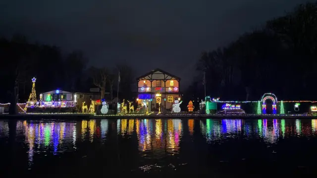Una casa con múltiples elementos de iluminación de estilo festivo, incluidos muñecos de nieve, renos y árboles, que en el exterior brillan y se reflejan en un cuerpo de agua.