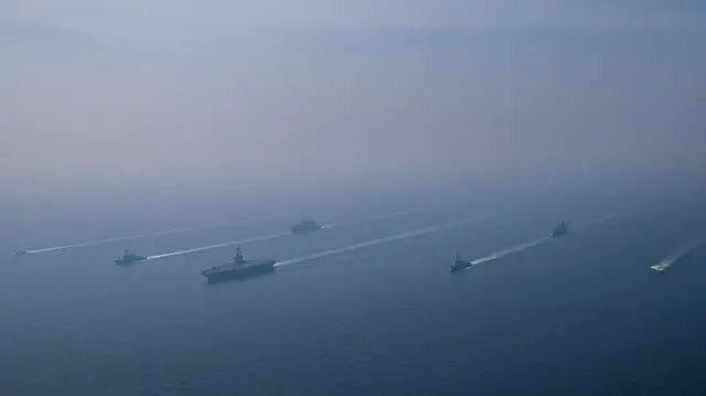 نمای هوایی از گروه ضربتی ناو هواپیمابر یو‌اس‌اس آبراهام لینکلن در حال عملیات در دریای عرب، همراه با دو ناو تأمین لجستیک نظامی و دو فروند کشتی گارد ساحلی ایالات متحده، در حالی که جنگنده‌های ناوگان هوایی شماره نه بالای سر آنها به انجام پروازهای عملیاتی مشغول بودند، در تاریخ ۱۷ بهمن ۱۴۰۴.
