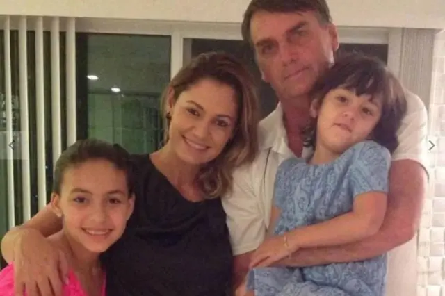 Jair Bolsonaro com Michelle Bolsonaro e as duas filhas. Michelle e Letícia sorriem olhando para a câmera, enquanto Bolsonaro está sério e Laura, em seu colo, morde o lábio inferior