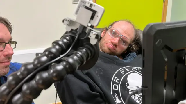 Primer plano de un equipo de tecnología asistencial con brazos flexibles y un dispositivo montado cerca de una persona que viste una sudadera negra con capucha con un logotipo circular y el texto "DR PEACE". En primer plano se ve la pantalla de una tableta y al fondo una pared de color verde brillante.