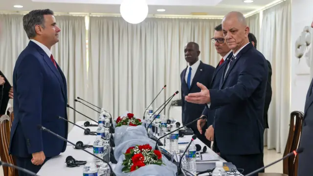 Uma fila de homens de terno em pé ao redor de uma longa mesa com garrafas de água de plástico e microfones finos, em frente a um homem que está sozinho à mesa