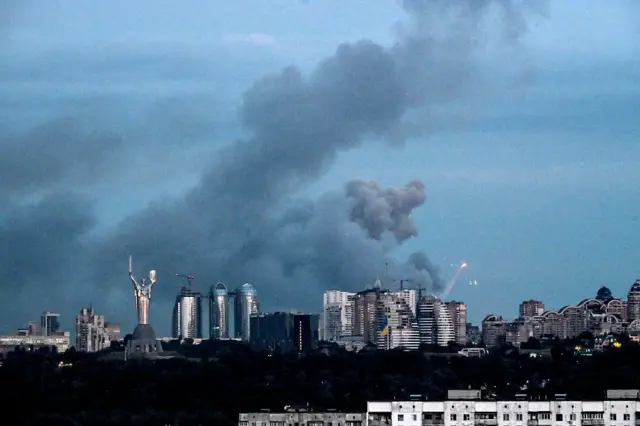 Київ під час російської атаки — над житловими кварталами та діловим центром міста здіймаються густі стовпи диму після вибухів. На передньому плані видно монумент «Батьківщина-Мати», а поруч із хмарочосами — спалахи та сліди роботи систем протиповітряної оборони.