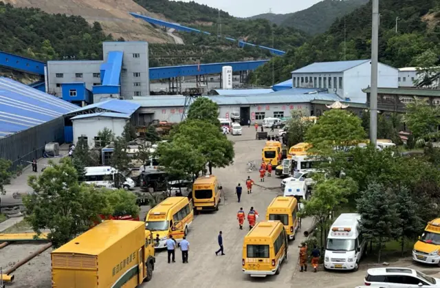 Sarı renkli kurtarma araçlar ve kırmızı giyen görevlileri uzaktan görüyoruz. Madenin girişi ve kurulu olduğu dağlık alan da arkada
