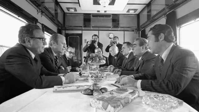 Генри Киссинжер (алдынкы планда сол жакта). АКШнын президенти Джералд Форд (Киссинжердин жанында) жана америкалык делегациянын башка мүчөлөрү 1974-жылы Советтер Союзун аралап бара жаткан поездде КПССтин Борбордук Комитетинин Генералдык секретары Леонид Брежнев, СССРдин тышкы иштер министри Андрей Громыко жана СССРдин АКШдагы элчиси Анатолий Добрынин менен маектешүүдө.