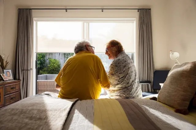 Dois idosos conversam em uma cama com um cobertor amarelo, em frente a uma janela onde o sol brilha.