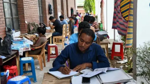 কয়েকজন তরুণ সারিবদ্ধভাবে রাখা চেয়ার-টেবিলে বসে পড়ছেন