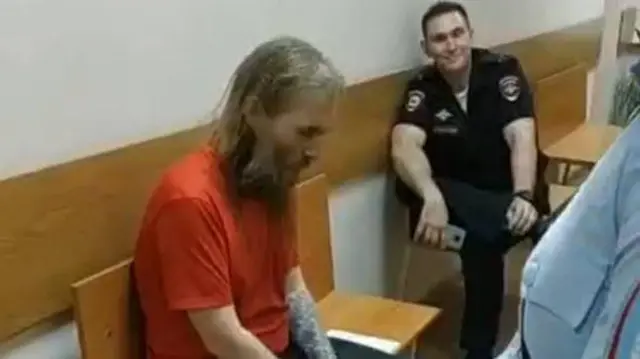 Березиков сидит перед залом суда 31 мая, рядом довольный конвоир