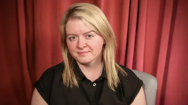 A woman with blonde hair in a black top sits on a chair in front of a red curtain