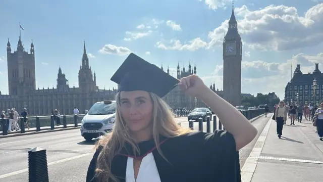 Melys stands in front of a London skyline with Big Ben and the Houses of Parliament behind her. She wears a cap and gown, with her hand holding the tip of her graduation cap.