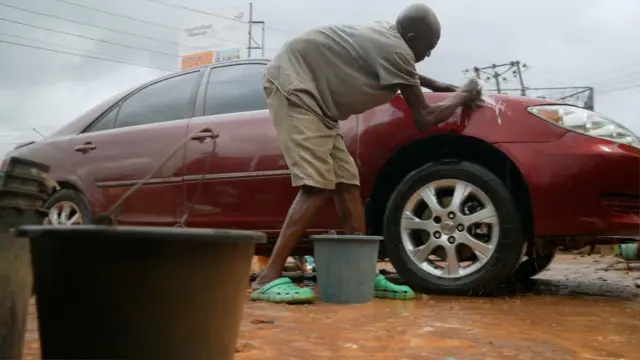 Ọkan ninu awọn baba naa wọ asọ ati sokoto penpe alawọ eeru, o wọ bata rọba konko, to si n fi ọsẹ fọ mọto alawọ pupa kan.