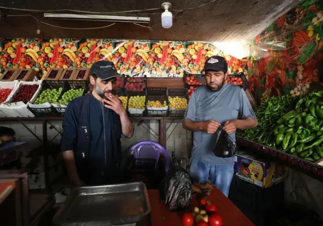 Dos hombres en el centro de una tienda de verduras, uno de ellos, el de la izquierda, vestido de negro, lleva una gorra en la cabeza y está fumando un cigarro. El otro, a la derecha, también lleva gorra en la cabeza y en la mano sostiene una bolsa negra, llena de verdura. Están rodeador de estantes llenos de pimientos, manzanas, tomates, cebollas. Delante de ellos, una mesa con una balanza y verdura.