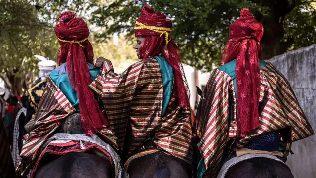 Three men in striped robes and turbans sit on horses, with their backs to the camera