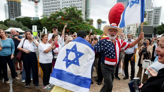 Uma pessoa enrolada na bandeira de Israel dança com outra vestida com uma camiseta estampada com a bandeira dos Estados Unidos em uma rua em Tel Aviv.