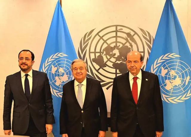 Nikos Hristodulidis, Antonio Guterres ve Ersin Tatar BM bayrakları önünde kameralara bakıyor.