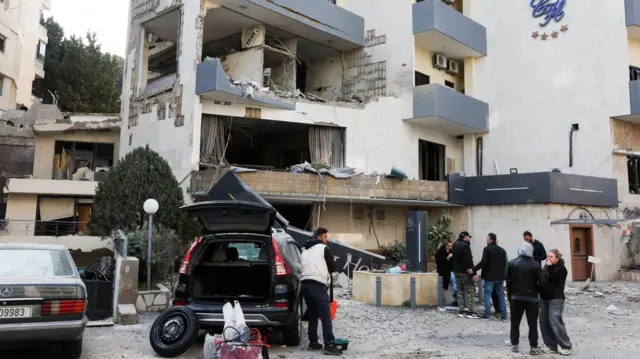 Several people stand outside a damaged building with its windows blown out. 