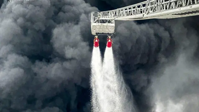 Gust, tamnosivi dim kulja iz naftnog čvorišta Brodi dok vatrogasci gase nevidljivi požar sa dva creva sa produženih merdevina.