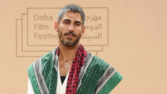 A photo of Kuwaiti‑American journalist Ahmed Shihab-Eldin, with the 2025 Doha Film Festival logo visible in the background. He has grey hair and a black and grey beard and is wearing a green, red and white patterned top as well as a necklace.