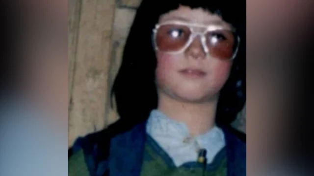 A close-up, slightly blurry shot of Zahra dressed as a boy. She has thick black chin-length hair with a heavy fringe, and wears aviator style sunglasses. She has a white shirt and a blue waistcoat.