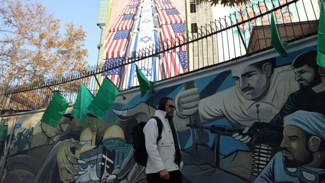 Мужчина стоит на фоне граффити, прославляющего иранских военных, а на заднем фоне здание с большим билбордом с гробами, обтянутыми израильскими и американскими флагами и с угрозами в адрес американских и израильских военных на иврите и на английском.