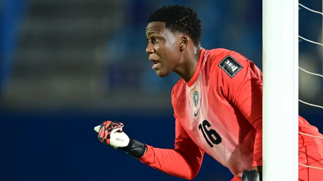 Chiamaka Nnadozie, vêtue de gants de gardienne et d'un maillot orange vif arborant le numéro 16 noir sur la poitrine, pointe son pouce droit devant elle tout en cherchant à organiser une défense qui se trouve hors champ. Elle se tient près d'un poteau de but blanc, dont le filet est visible sur le côté de la photo.