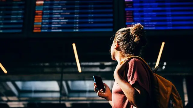Mulher com celular olha para o quadro de partidas e chegadas de um aeroporto