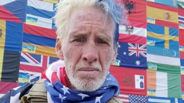 Selfie show Ryan W. Routh in military bulletproof vest wearing red white and blue t-shirt and bandana around im neck wit wall of flags of di world behind am