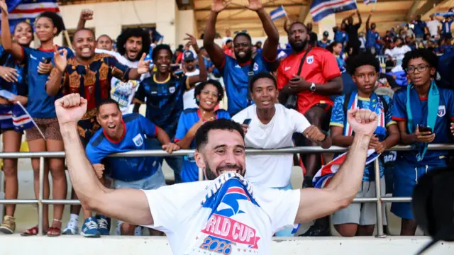 Roberto Lopes, visible de la poitrine vers le haut au premier plan de l'image, tient ses bras en l'air alors qu'il célèbre la qualification du Cap-Vert pour la Coupe du monde 2026. Il porte un tee-shirt blanc avec l'inscription World Cup 2026 dessus, qu'il soulève en le mordant entre ses dents. Douze supporters capverdiens joyeux, portant pour la plupart des maillots bleus de l'équipe, sont vus dans les gradins derrière lui, tandis que d'autres personnes et des drapeaux du Cap-Vert sont vus au loin