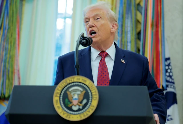 Trump falando ao microfone diante de palanque de presidente dos EUA; ele está dentro de sala