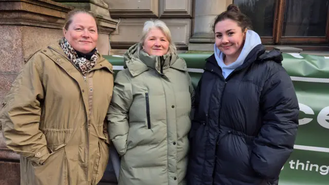 Tres mujeres de pie frente al Ayuntamiento de Glasgow. Una lleva una chaqueta color canela, una bufanda con estampado de leopardo y tiene el pelo rubio y castaño. Otra lleva un abrigo verde claro y el pelo rubio. La tercera lleva un abrigo de invierno grande negro/azul oscuro y el pelo oscuro.