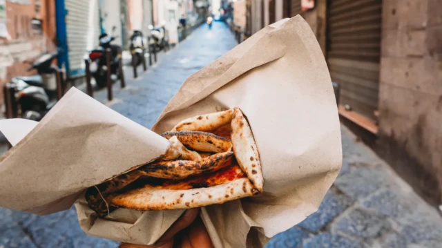 O persoană se plimbă pe o stradă din Italia, ținând în mână felii de pizza învelite în hârtie.
