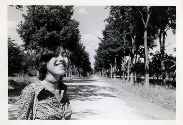 Mariona jovem em foto em preto e branco. Ela está em uma estrada cercada por árvores e sorri.