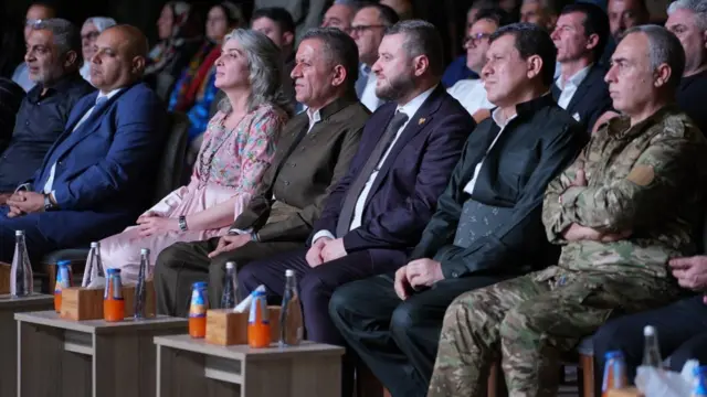 Abdi sağdan ikinci, sağında askeri kamuflaj giyen biri ve sol tarafından resmi giyimli insanlar var.
