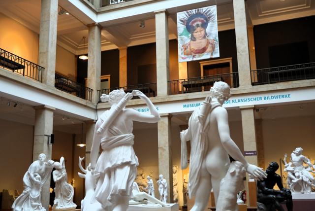 Colagem digital com imagem de Miranha no alto de pé direito de museu; em primeiro plano, vê-se esculturas clássicas