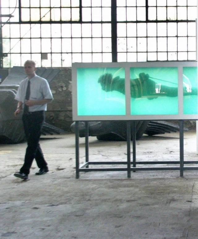 Dans une pièce vitrée, la sculpture, enfermée dans une boîte, a les mains et les pieds liés. Un homme passe à côté de l'œuvre.