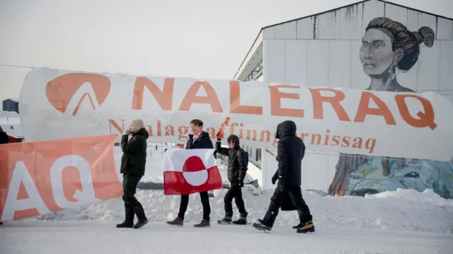 Тепло одетые люди идут по заснеженной дороге, у одного из них в руках бело-красный флаг Гренландии. На фоне — растяжка политической партии Naleraq.