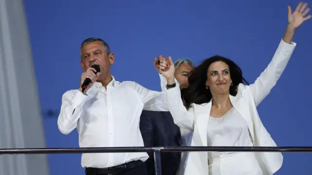 Özgür Özel miting otobüsünde elinde mikrofon diğer eliyle Rızvanoğlu'nun elini tutuyor. Rızvaoğlu beyaz giyinmiş halkı selamlıyor.