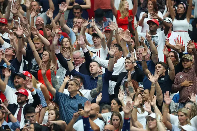 Un grupo de personas de diferentes edades y razas alzan los brazos en alabanza durante el evento conmemorativo de Charlie Kirk